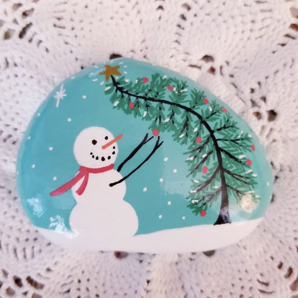 Snowman decorating a Christmas tree rock painting